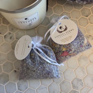 May include: Two small, sheer white organza sachets filled with dried purple lavender buds and colorful flower petals, each featuring a round white paper tag with the text Cavalli Farms Lavender, and a large white candle with a matching label.