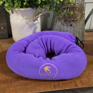 May include: A purple travel neck pillow with a drawstring closure. The pillow is made of a soft, ribbed fabric and features a gold logo with a horse and the text "Cavalli". The pillow is designed to provide support and comfort during travel.