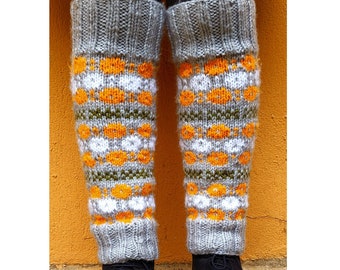 May include: Gray knitted leg warmers with orange, white, and green geometric patterns. The leg warmers are worn over black leggings and paired with black boots. The background is a solid orange wall.