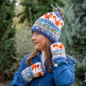 Può includere: Un cappello di lana blu, bianco, arancione e verde con un pon pon in cima. Il cappello ha un motivo di ghiande e volpi. Il cappello è indossato da una persona che indossa una camicia di jeans blu.