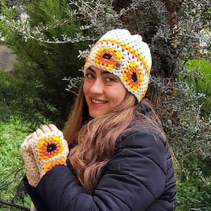 May include: A white crocheted hat with a sunflower design in orange, yellow, and black. The hat is worn by a person with long brown hair. The person is also wearing matching crocheted fingerless gloves with a sunflower design.