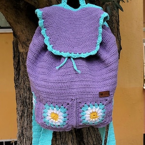 Può includere: Zaino in crochet viola con spalline turchesi e dettagli a forma di margherita. Lo zaino ha una chiusura a coulisse e una piccola etichetta marrone con il testo "Hand Made".