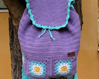 May include: Purple crocheted backpack with turquoise straps and daisy-shaped details. The backpack has a drawstring closure and a small brown label with the text "Hand Made".