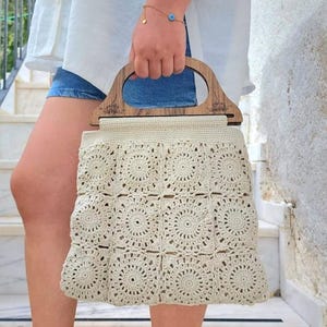 May include: A beige crocheted tote bag with a wooden handle. The bag is made of a square pattern with circular designs.