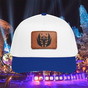 Op de afbeelding: Witte en blauwe baseballcap met een bruin leren patch met een zwart grafisch ontwerp. De cap heeft een gebogen klep en een blauwe klep. Het ontwerp op de patch lijkt een gestileerde vogel of wezen te zijn.