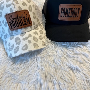 May include: Two trucker hats, one black with a brown leather patch that says "SOMEBODY" and one white with a brown leather patch that says "Somebody's PROBLEM".