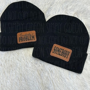 May include: Two black knit beanies with brown leather patches. One patch says "Somebody's Problem" and the other says "Somebody".