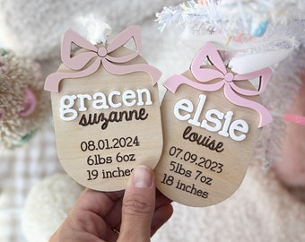 Puede incluir: Dos adornos de madera rosa y blanco con lazos rosas. Los adornos están personalizados con los nombres "Gracen Suzanne" y "Elsie Louise" e incluyen las fechas de nacimiento, los pesos y las longitudes.