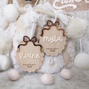 May include: Two personalized wooden Easter ornaments with gold bows and the names "Sloane" and "Myla" with the year "2025" on each. The ornaments are hanging from a white fluffy basket with a bunny doll and a banner that says "Happy Easter".