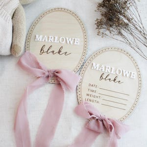 May include: Two oval wooden plaques with scalloped edges, each featuring the name MARLOWE blake in white and brown lettering. One plaque includes blank lines for date, time, weight, and length. Both are adorned with dusty pink silk ribbons.
