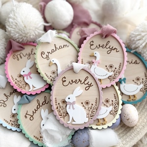 May include: Wooden Easter-themed name tags with scalloped edges in various pastel colors. Each tag features a name, a laser-cut bunny or duck, and floral details. Ribbons and pom-poms add to the festive decor.