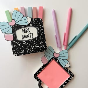 Op de afbeelding: Een bureau-organizer met een thema van een zwart-wit compositieboek met de naam "MRS. MONTZ" op de voorkant. Een roze vierkant notitieblok en verschillende pastelkleurige pennen en markeerstiften zijn ook zichtbaar.