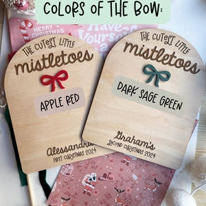Puede incluir: Dos adornos de madera con forma de arco con el texto "The Cutest Little Mistletoes" y las palabras "Apple Red" y "Dark Sage Green" escritas debajo. Los adornos est&aacute;n decorados con un lazo rojo y un lazo verde.