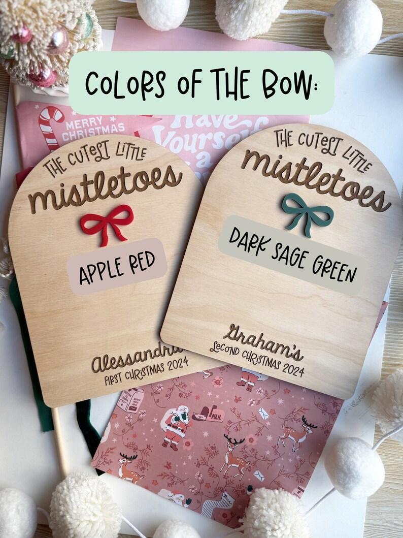 Puede incluir: Dos adornos de madera con forma de arco con el texto "The Cutest Little Mistletoes" y las palabras "Apple Red" y "Dark Sage Green" escritas debajo. Los adornos est&aacute;n decorados con un lazo rojo y un lazo verde.