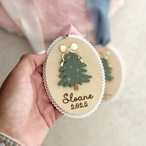 May include: Wooden Christmas ornament with a green tree design, gold bow, and the name "Sloane" and the year "2025". The ornament has a scalloped edge and a pink ribbon for hanging.