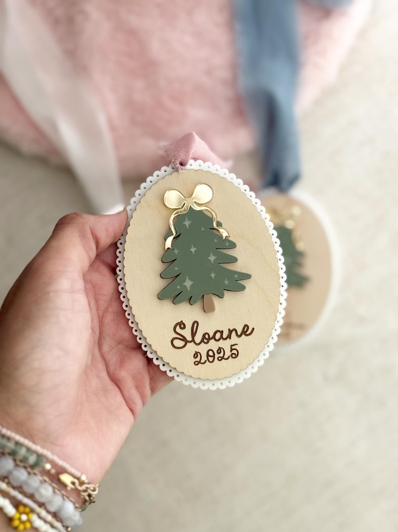 May include: Wooden Christmas ornament with a green tree design, gold bow, and the name "Sloane" and the year "2025". The ornament has a scalloped edge and a pink ribbon for hanging.