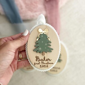 May include: Wooden Christmas ornament with a green tree design and gold bow. The ornament has the text "Baker first Christmas 2025". The ornament is oval-shaped with a scalloped edge and a white ribbon for hanging.