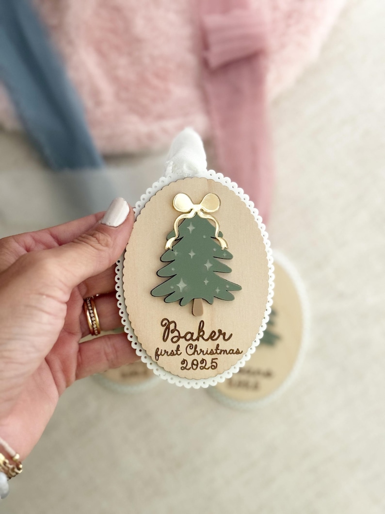 May include: Wooden Christmas ornament with a green tree design and gold bow. The ornament has the text "Baker first Christmas 2025". The ornament is oval-shaped with a scalloped edge and a white ribbon for hanging.