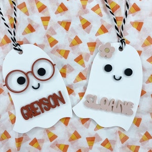 May include: Two white ghost ornaments with black eyes and mouths. One ghost has red glasses and the name Greyson in brown letters. The other ghost has a pink flower and the name Sloane in pink letters.