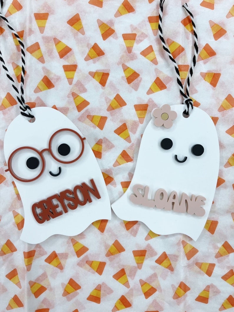 May include: Two white ghost ornaments with black eyes and mouths. One ghost has red glasses and the name Greyson in brown letters. The other ghost has a pink flower and the name Sloane in pink letters.