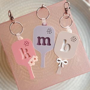 Könnte beinhalten: Drei rosa, lila und beigefarbene paddelförmige Schlüsselanhänger mit den Buchstaben "k", "m" und "b" darauf. Jeder Schlüsselanhänger hat eine kleine weiße Schleife und einen kleinen weißen Kreis mit einem schwarzen Punkt in der Mitte.
