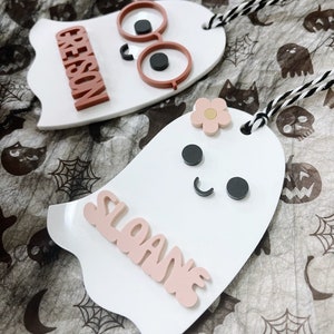 May include: Two white ghost-shaped ornaments with names "Sloane" and "Greyson" written in pink and brown letters. The ghost with the name "Sloane" has a pink flower and black eyes. The ghost with the name "Greyson" has brown glasses and black eyes.