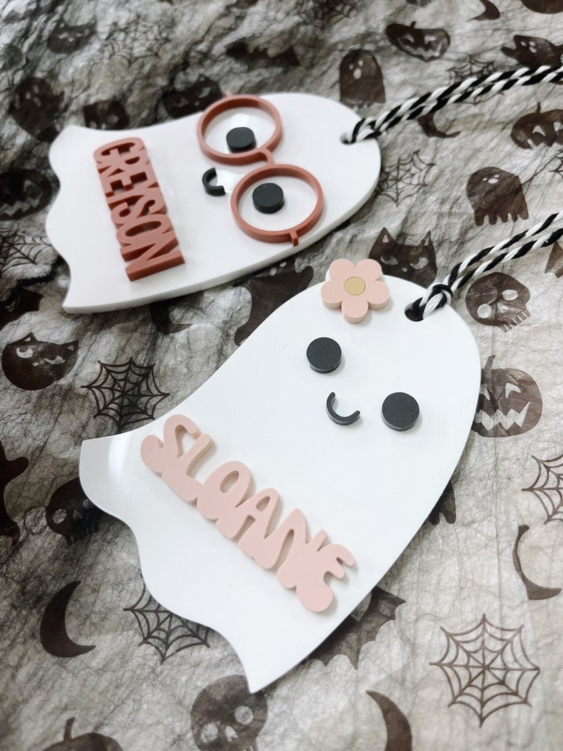 May include: Two white ghost-shaped ornaments with names "Sloane" and "Greyson" written in pink and brown letters. The ghost with the name "Sloane" has a pink flower and black eyes. The ghost with the name "Greyson" has brown glasses and black eyes.