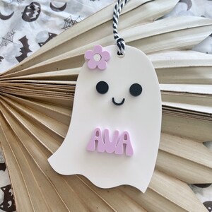 May include: A white ghost-shaped ornament with a pink flower, black eyes, and a smiling mouth. The ghost has the name "AVA" written in pink letters.