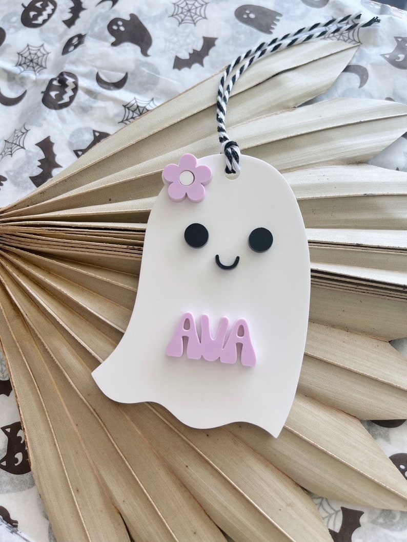 May include: A white ghost-shaped ornament with a pink flower, black eyes, and a smiling mouth. The ghost has the name "AVA" written in pink letters.
