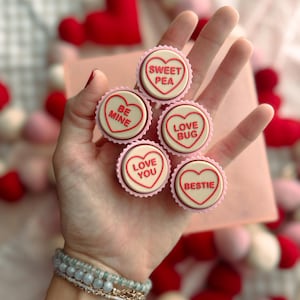 May include: A hand holding five heart-shaped candies with messages like "Sweet Pea" and "Be Mine" in red lettering. The candies are in pink cupcake liners. Valentine's Day theme.