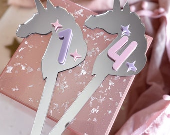 May include: Two silver unicorn-shaped cake toppers with pink and purple numbers 1 and 4. The unicorn toppers are on a pink and white glittery surface.