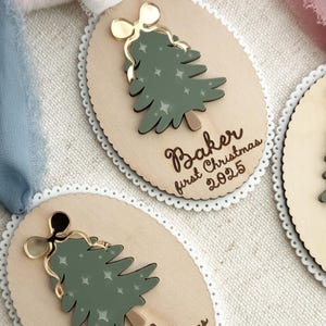 May include: Wooden Christmas tree ornaments with a green tree design, gold bow, and the text "Baker first Christmas 2025" and "Eli first Christmas 2025". The ornaments have a scalloped edge and a ribbon for hanging.