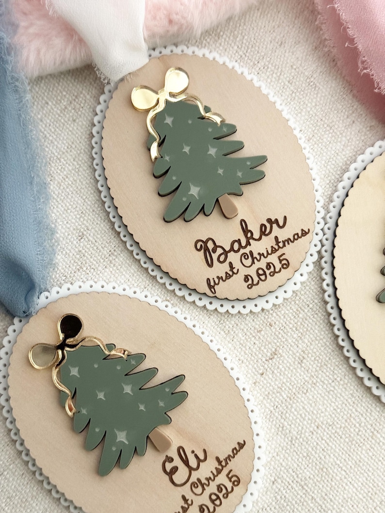May include: Wooden Christmas tree ornaments with a green tree design, gold bow, and the text "Baker first Christmas 2025" and "Eli first Christmas 2025". The ornaments have a scalloped edge and a ribbon for hanging.