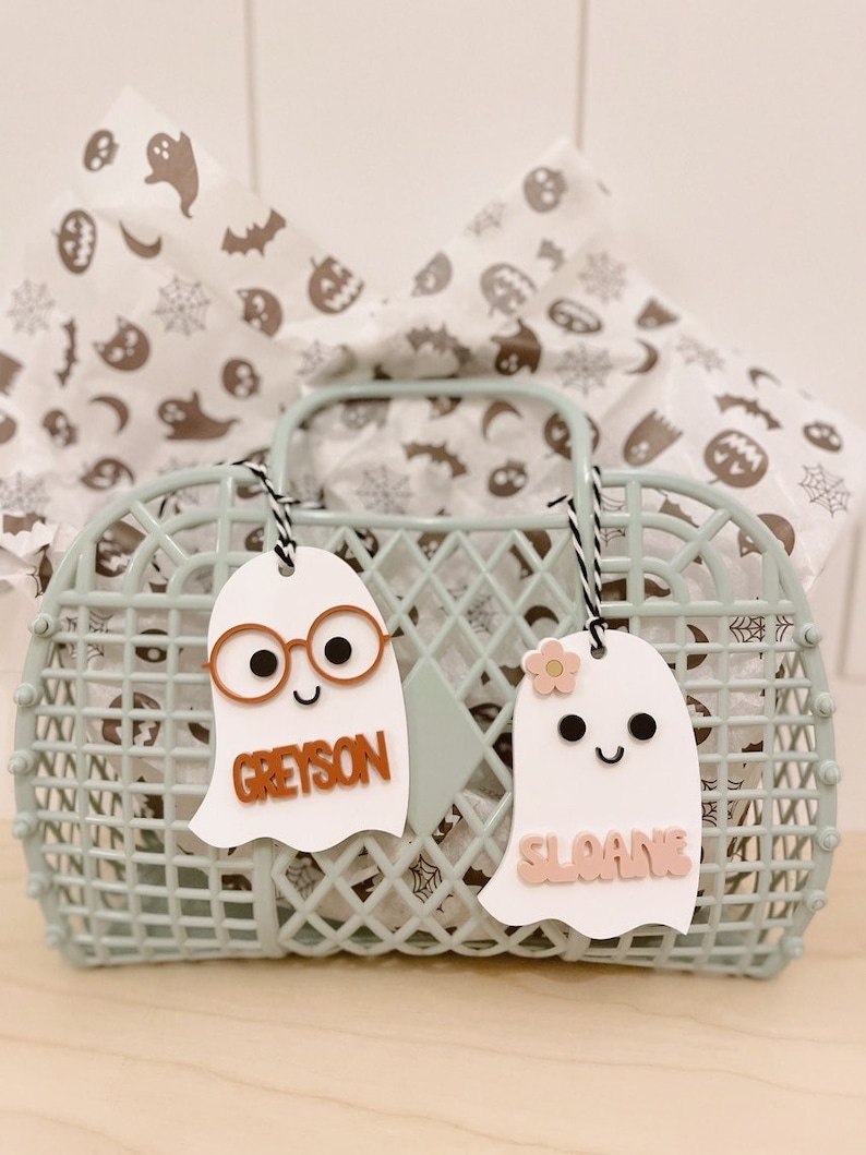 May include: Two white ghost-shaped Halloween gift tags with names "Greyson" and "Sloane" written in orange and pink letters. The tags are hanging from a light blue wire basket filled with white tissue paper with a Halloween pattern.