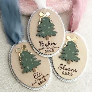 May include: Three wooden Christmas tree ornaments with gold bows and green trees. Each ornament has a name and the year 2025. The ornaments are attached to ribbons in blue, white, and pink.