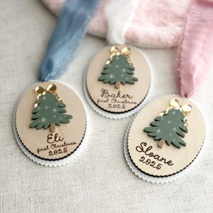 May include: Three wooden Christmas ornaments with a green tree design and gold bow. Each ornament has a name and the year 2025. The ornaments are on a white scalloped edge with a ribbon.