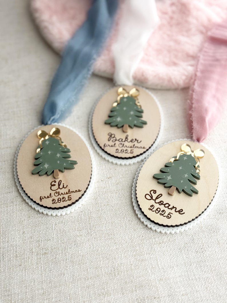 May include: Three wooden Christmas ornaments with a green tree design and gold bow. Each ornament has a name and the year 2025. The ornaments are on a white scalloped edge with a ribbon.