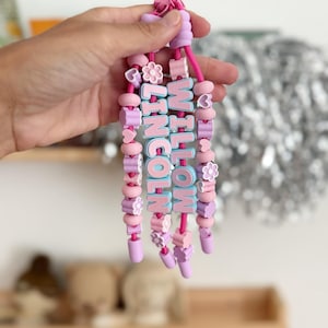 May include: A set of three beaded keychains on pink cords, featuring light blue letter beads spelling WILLOW and LINCOLN. The strands are decorated with various pastel-colored silicone beads in shapes of hearts, flowers, and circles in pink and purple.