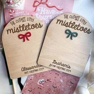 Puede incluir: Dos letreros de madera con el texto "The Cutest Little Mistletoes" y un lazo rojo y verde. Los letreros est&aacute;n personalizados con los nombres "Alessandra" y "Graham's" y los a&ntilde;os "First Christmas 2024" y "Second Christmas 2024".