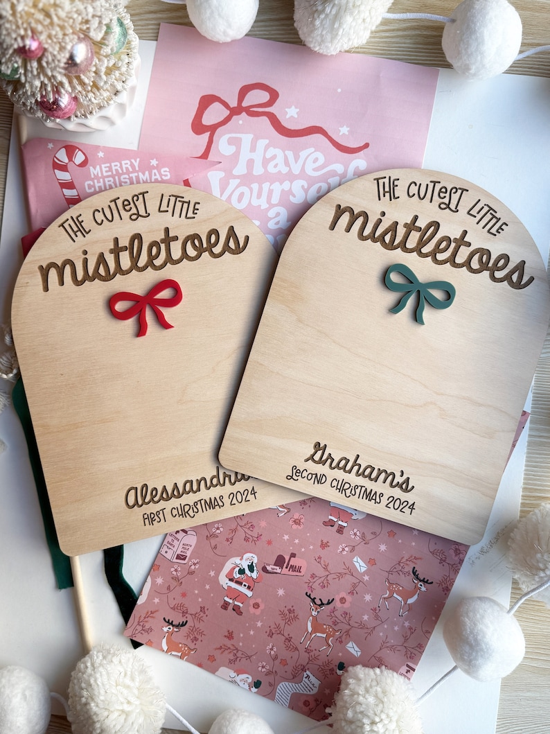 Puede incluir: Dos letreros de madera con el texto "The Cutest Little Mistletoes" y un lazo rojo y verde. Los letreros est&aacute;n personalizados con los nombres "Alessandra" y "Graham's" y los a&ntilde;os "First Christmas 2024" y "Second Christmas 2024".