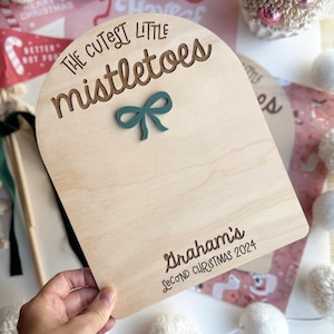 Puede incluir: Un letrero de madera con la parte superior arqueada, decorado con el texto "The Cutest Little Mistletoes" y un lazo verde. El letrero est&aacute; personalizado con el nombre "Graham's" y el texto "Second Christmas 2024".