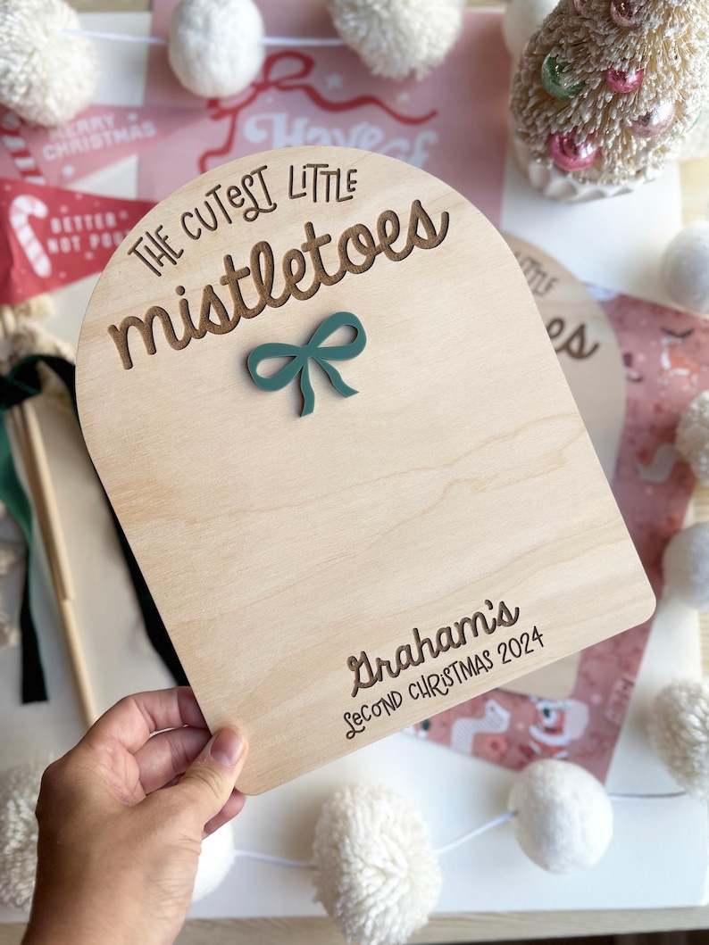 Puede incluir: Un letrero de madera con la parte superior arqueada, decorado con el texto "The Cutest Little Mistletoes" y un lazo verde. El letrero est&aacute; personalizado con el nombre "Graham's" y el texto "Second Christmas 2024".