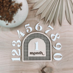 May include: Wooden arch with a woven pattern and a white number 1 in the center. The arch is surrounded by white numbers 1 through 9. The text "1 month" is below the number 1.