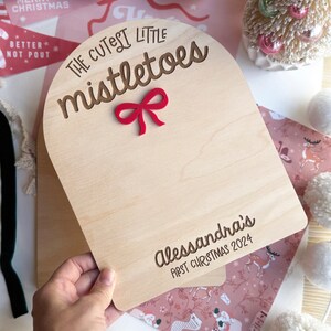 Puede incluir: Un letrero de madera con un lazo rojo y el texto "The Cutest Little Mistletoes" y "Alessandra's First Christmas 2024".