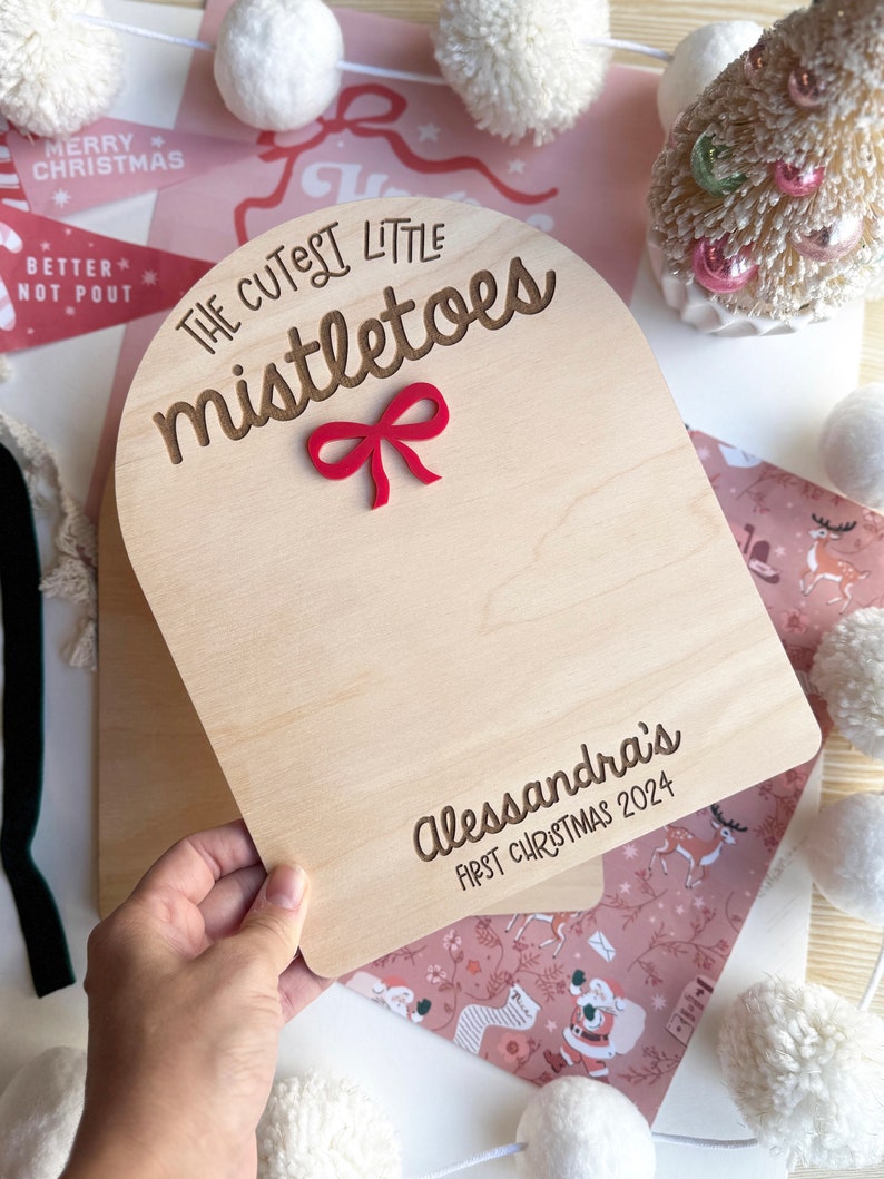 Puede incluir: Un letrero de madera con un lazo rojo y el texto "The Cutest Little Mistletoes" y "Alessandra's First Christmas 2024".