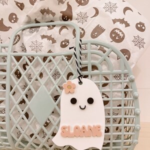 May include: A white ghost-shaped gift tag with a pink flower, black eyes, and a smiling mouth. The tag has the name "SLOANE" written in pink letters.