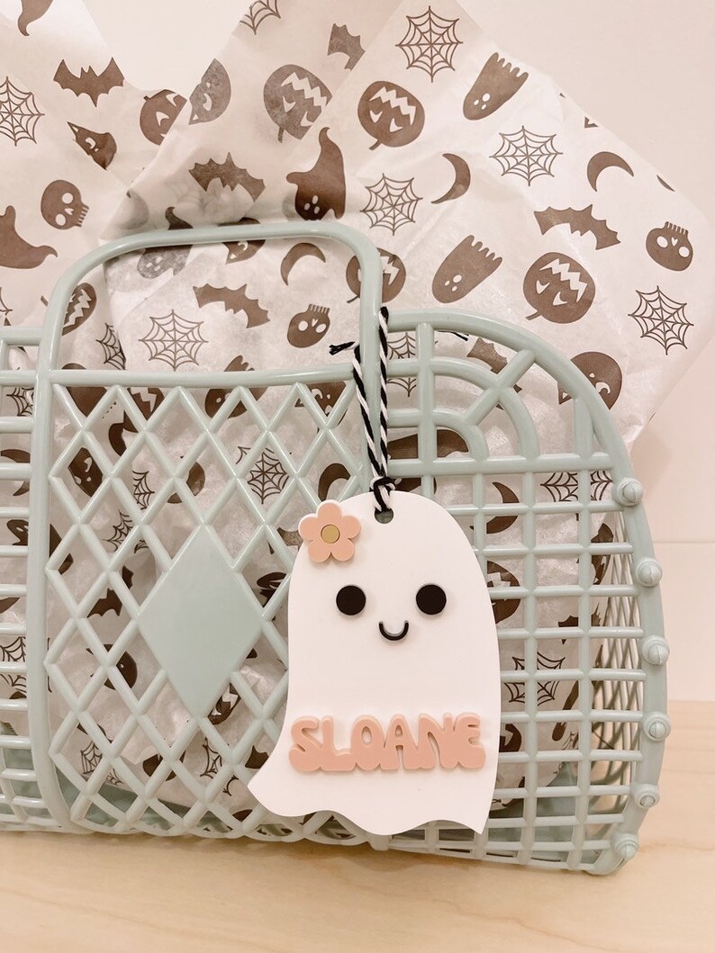 May include: A white ghost-shaped gift tag with a pink flower, black eyes, and a smiling mouth. The tag has the name "SLOANE" written in pink letters.