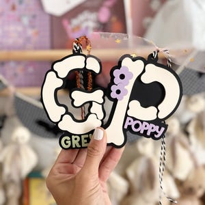 May include: Two Halloween-themed ornaments, each with a black and bone-colored design. One ornament spells out "GREY" and the other "POPPY", with purple flower accents. Both are held together with a black and white string.