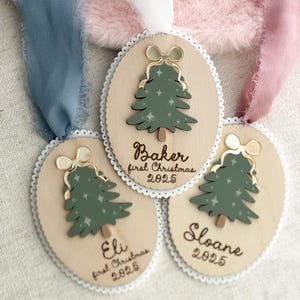May include: Wooden Christmas tree ornaments with a green tree design, gold bow, and white lace trim. Each ornament has a name and the year 2025. Ribbons in blue, pink, and white are attached to the ornaments.