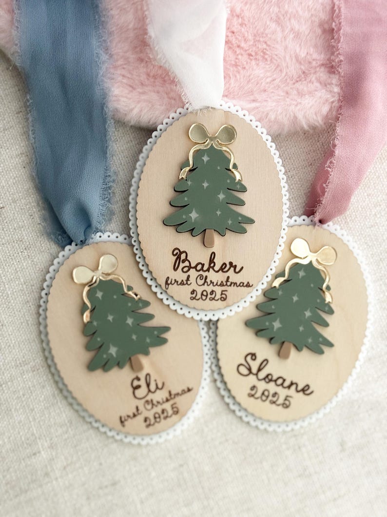 May include: Wooden Christmas tree ornaments with a green tree design, gold bow, and white lace trim. Each ornament has a name and the year 2025. Ribbons in blue, pink, and white are attached to the ornaments.